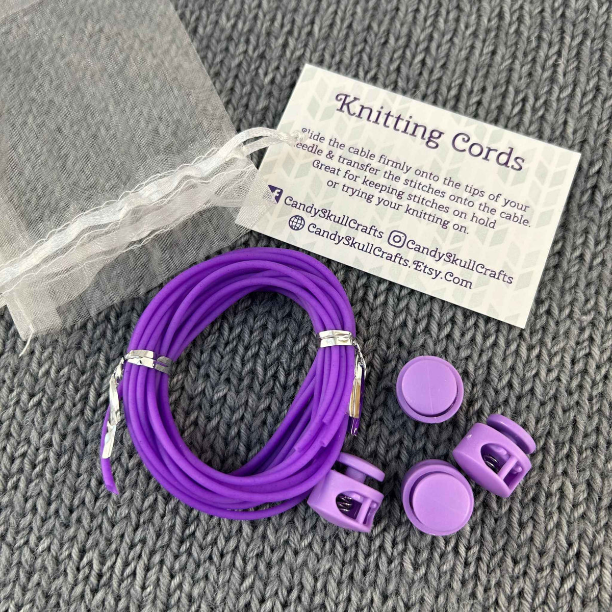 Purple cable knitting cords with matching stoppers and a small mesh bag, arranged on a gray knitted blanket beside a printed card that reads 'Knitting Cords' and shows the brand Candy Skull Crafts.
