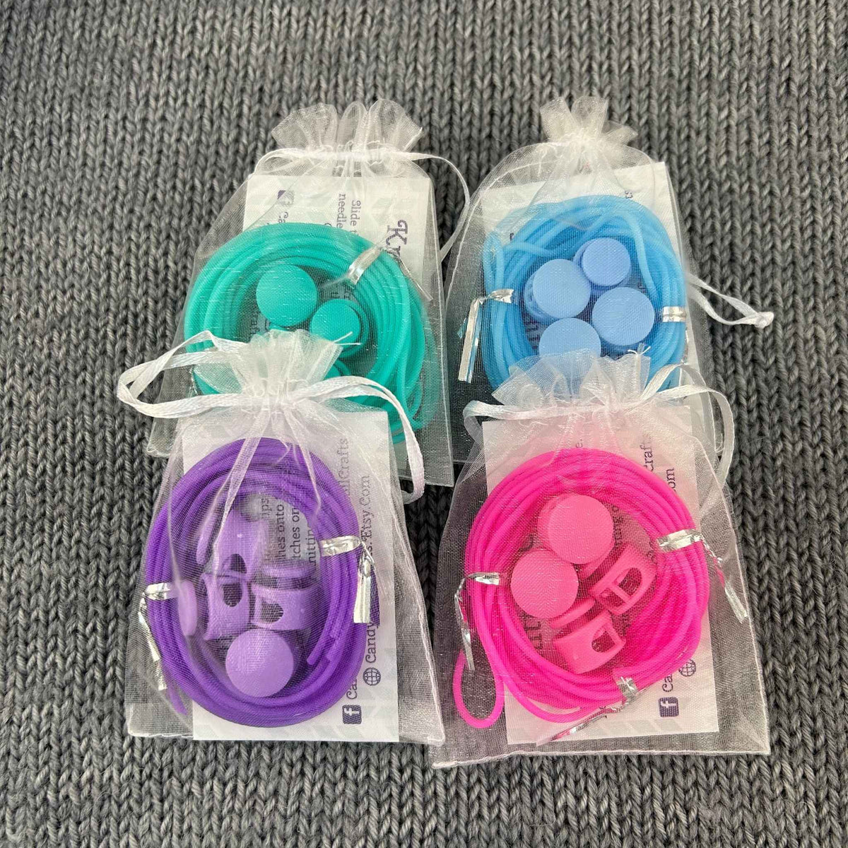 Four small clear drawstring pouches containing colorful silicone knitting stitch holder cords and 4 stoppers, arranged on a gray textured fabric background. The pouches contain sea green, blue, purple, and pink cords.