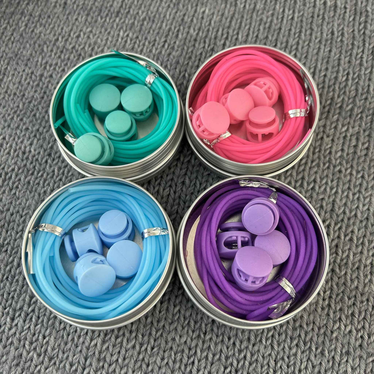 4 sets of knitting stitch holder try on cords with colour coordinating stoppers in metal tins.  Colours displayed are pink, blue, sea green, and purple
