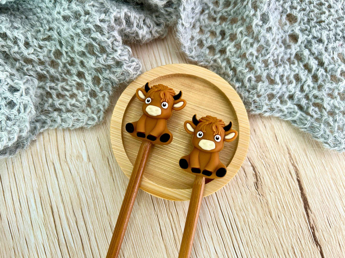 Two wooden knitting needles with cute cartoon cow-shaped stoppers, resting on a round bamboo plate over a light wood surface with a gray knitted blanket in the background.