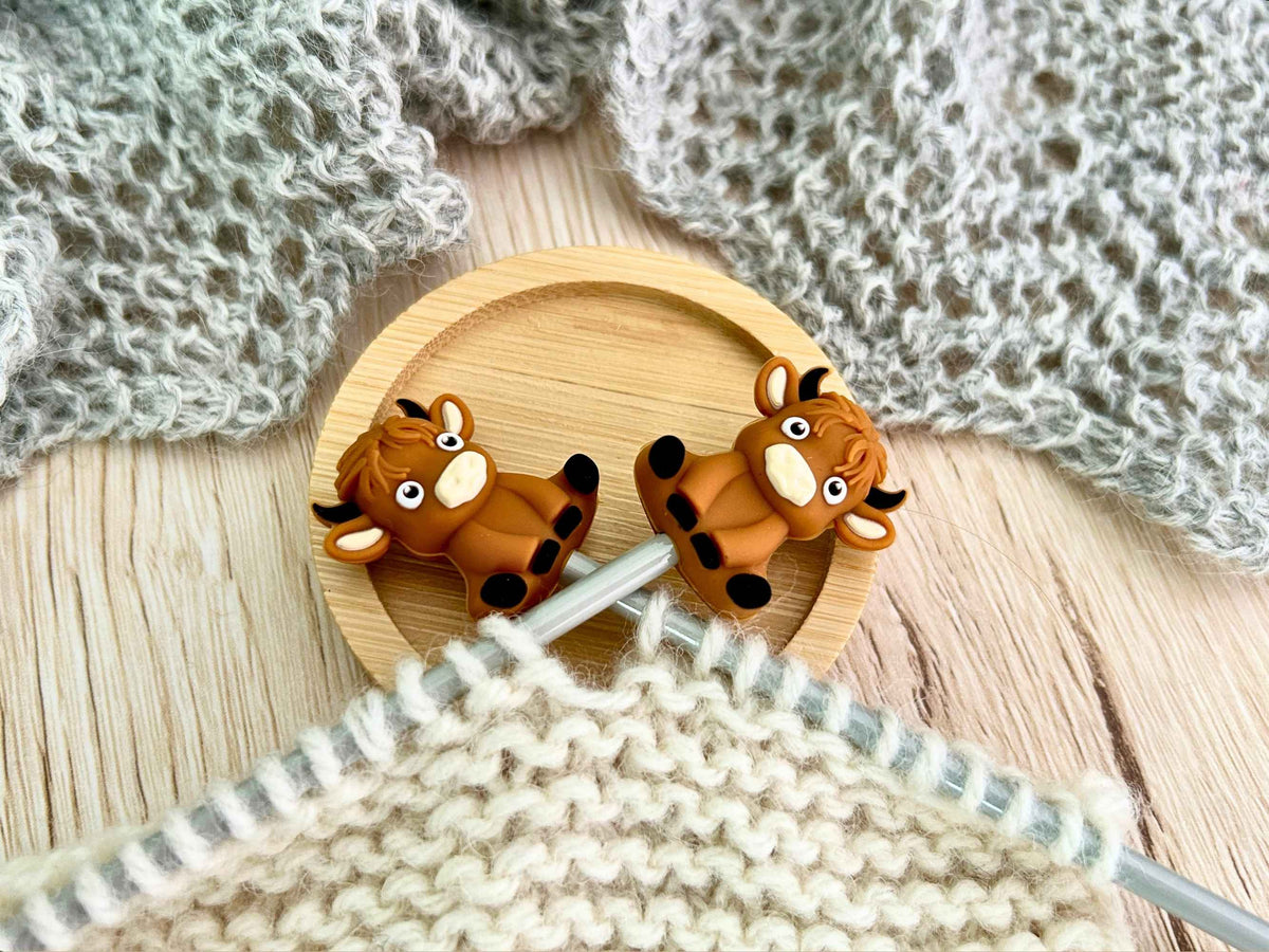 Two small brown cartoon highland cow knitting needle stoppers with black horns and white faces are attached to knitting needles to stop the cream knitting sliding off them The round wooden stand holding the needles sits on a light wood surface, with gray knitted fabric draped in the background.