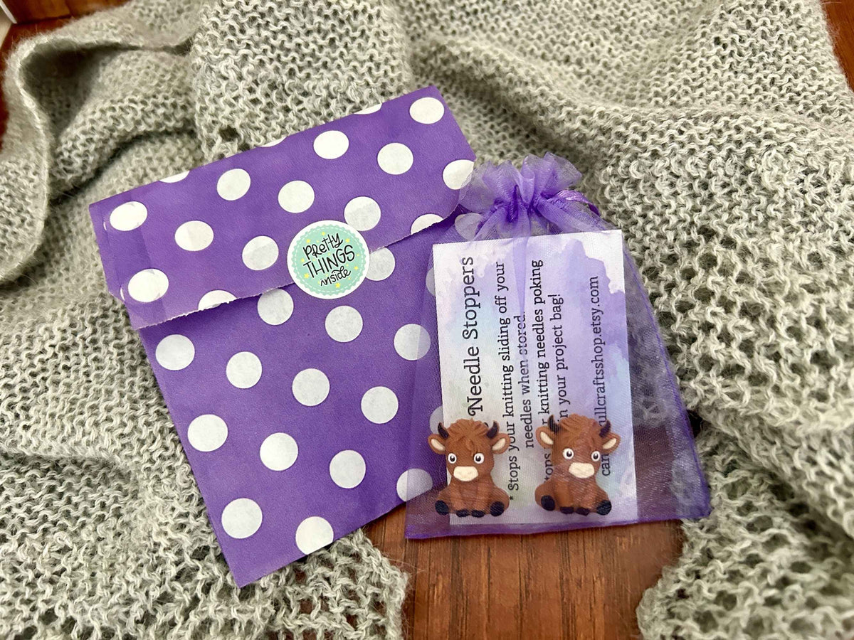 A purple and white polka dot paper bag, placed next to a sheer purple gift bag 'Needle Stoppers' card featuring two cartoon highland cows.