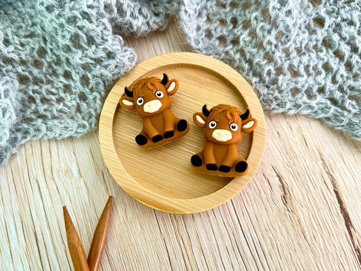 Two small brown cow-shaped knitting needle stoppers displayed on a round wooden tray, with a gray knitted textile and wooden knitting needles in the background.