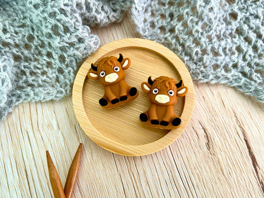 Two small brown cow-shaped knitting needle stoppers displayed on a round wooden tray, with a gray knitted textile and wooden knitting needles in the background.