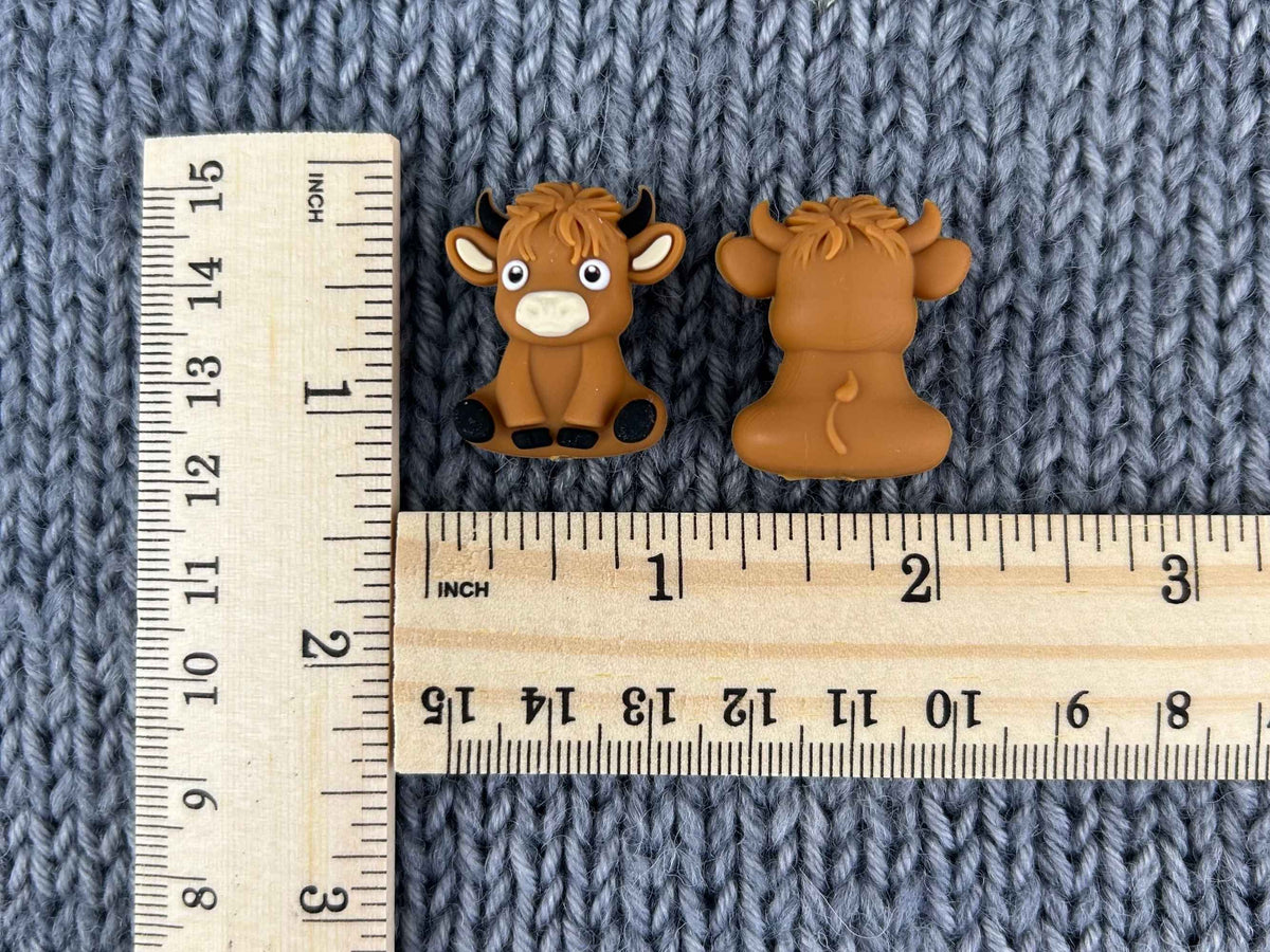 Two small brown cow-shaped knitting needle stoppers shown on a blue-gray knitted fabric background, with a wooden ruler measuring them as approx 24mm (0.94 inches) by 27mm (1.06 inches)