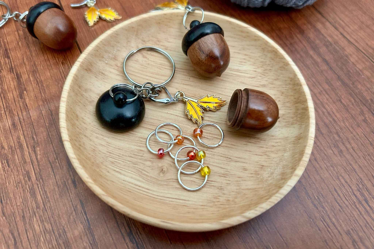 A dark coloured wooden acorn knitting stitch marker holder, opened to up reveal 6 beaded ring knitting stitch markers with red, orange, and yellow beads. There is a removable progress keeper n 2 yellow leaves. All displayed on a light wooden dish.