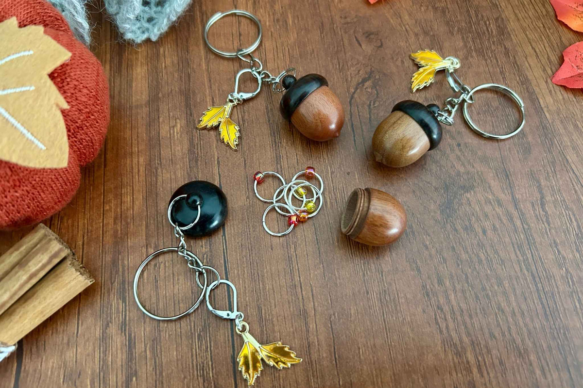 A dark coloured wooden acorn knitting stitch marker holder, opened to up reveal 6 beaded ring knitting stitch markers with red, orange, and yellow beads. There is a removable progress keeper n 2 yellow leaves. 