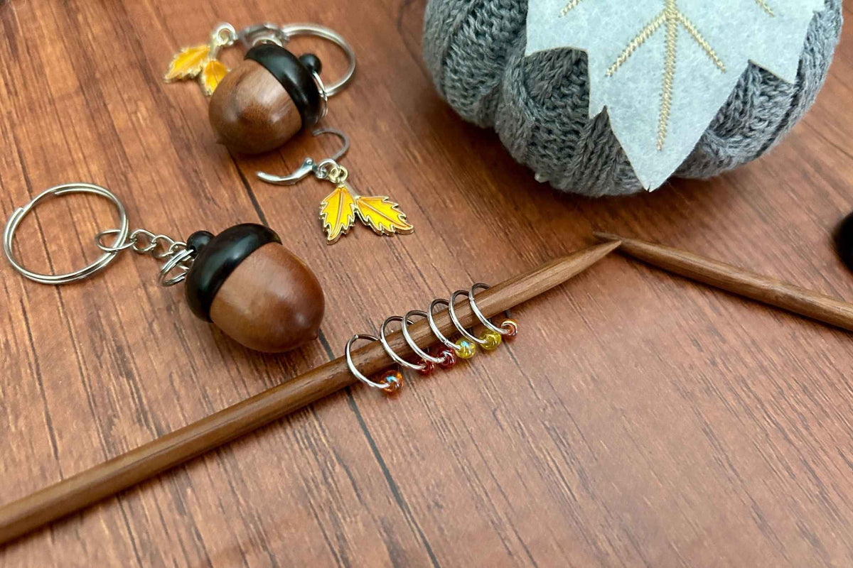 A dark coloured wooden acorn knitting stitch marker holder.  There is a removable progress keeper n 2 yellow leaves. the yellow, red, and orange beaded stitch markers are shown on a wooden knitting needle.
