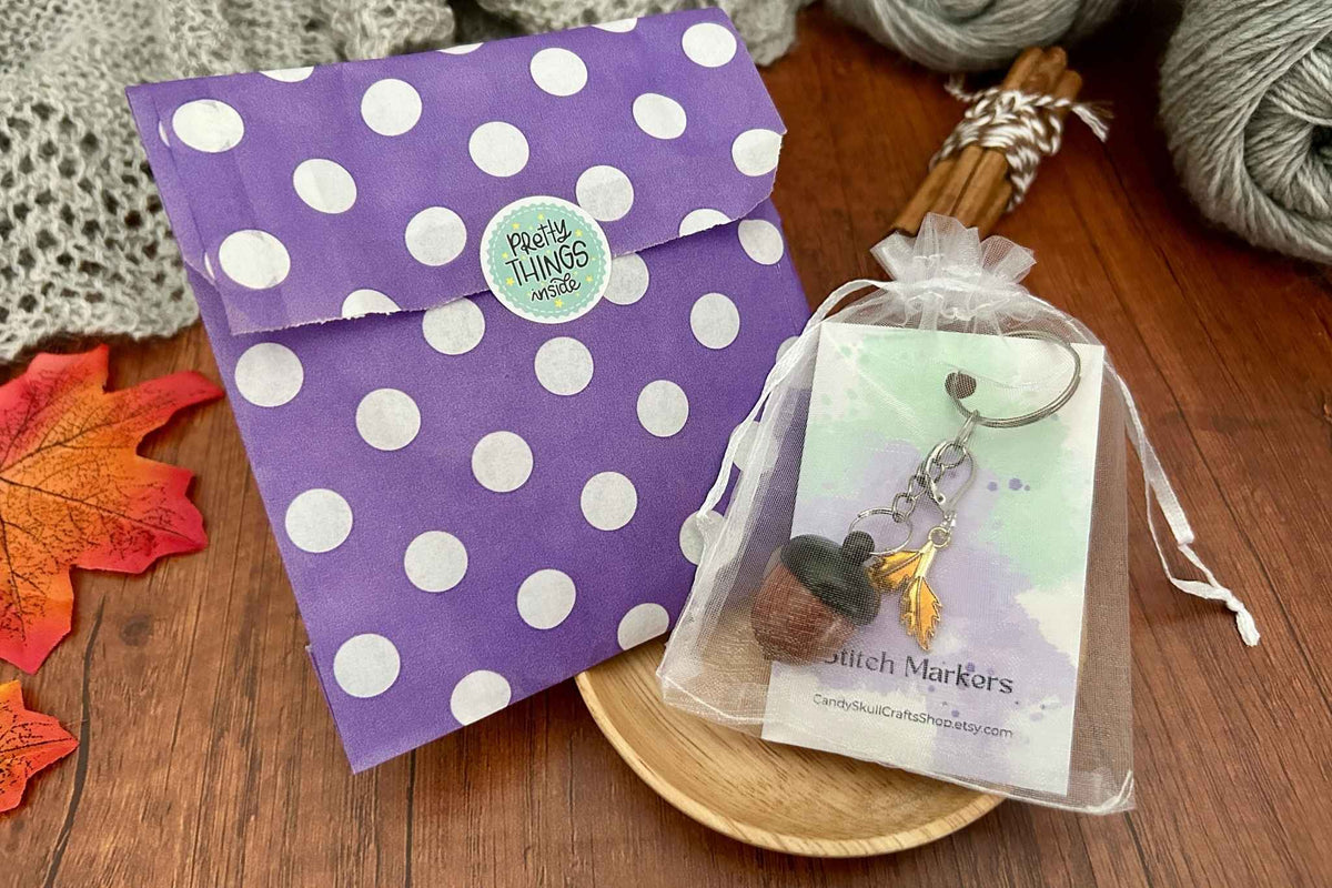 A dark coloured wooden acorn knitting stitch marker holder, in a mesh organza bag next to its packaging which is a purple and white polka dot bag with a sticker on which reads "pretty things inside" All displayed on a light wooden dish.