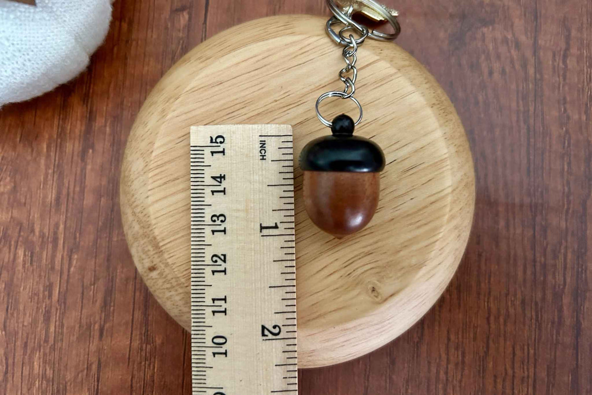 A wooden acorn knitting stitch marker holder on a wooden tray next to a ruler. The measurements are 1 inch x 0.7 inches