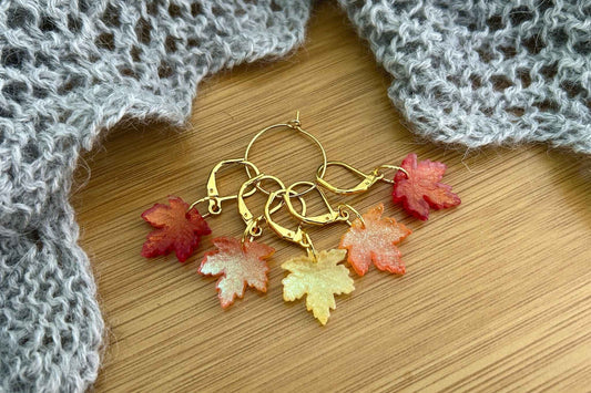 A set of 5 autumn themed maple lea knitting stitch markers or crochet progress keepers. 2 red shimmer, 2 orange shimmer, 1 yellow shimmer Set on a wooden background with a grey knitted shawl in the background