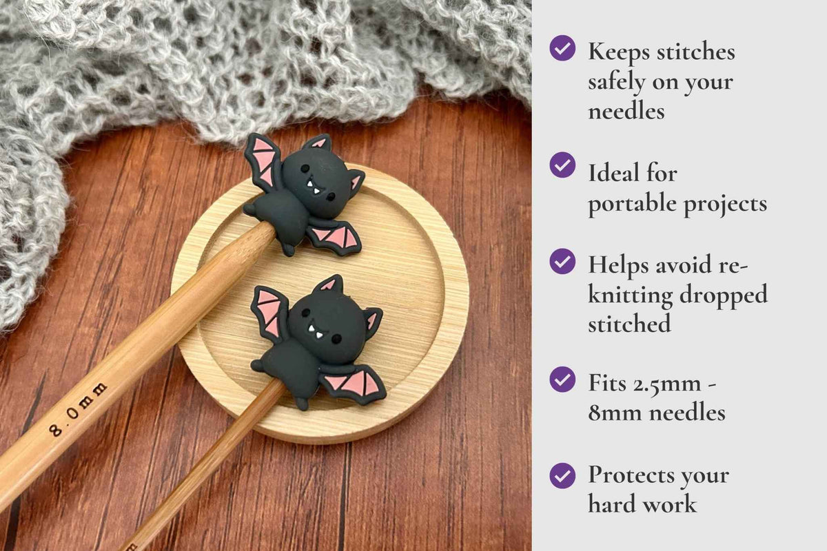 Two black bat-shaped stitch protectors with pink wings are shown on wooden knitting needles on a round wooden tray, with a knitted gray scarf in the background and a text panel listing benefits on the right. It reads Keeps stitches on needles. Ideal for portable projects. Helps avoid re-knitting dropped stitches. Fits mm to 2.5mm to 8mm needles 

