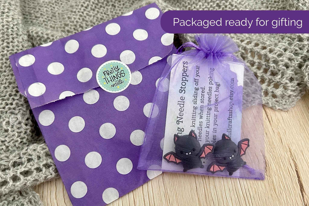 Purple and white polka dot gift packaging with a small purple organza bag containing black bat-shaped needle stoppers, shown on a wooden surface with a knitted blanket in the background. A purple banner reads 'Packaged ready for gifting'.