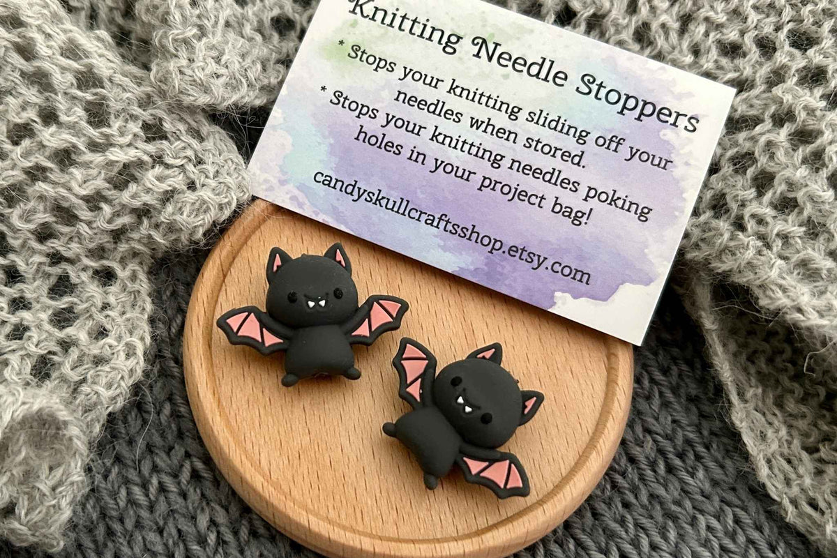 Two cute black bat-shaped knitting needle stoppers with pink wing interiors rest on a small round wooden tray, with a printed product card behind them on a gray knit blanket.