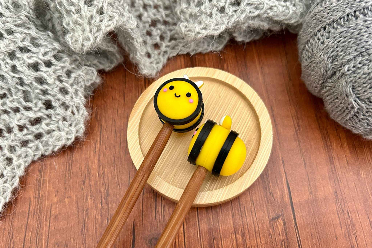 A pair of wooden knitting needles with cute yellow and black bee-shaped stoppers, resting on a small round wooden tray over a brown wooden table with gray knitted fabric in the background.