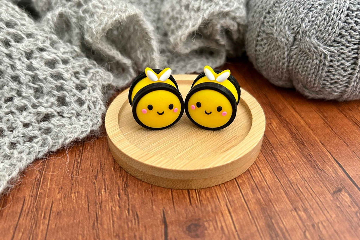 Two cute yellow and black bumblebee knitting needle stoppers with white accents and pink cheeks, displayed on a small round wooden coaster on a wooden table with a gray knit blanket in the background.