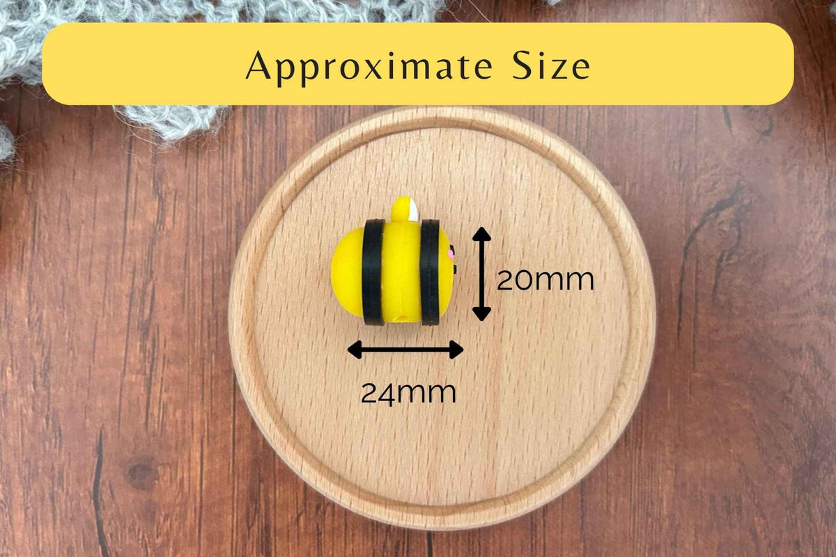 A small yellow and black bee-shaped knitting needle stopper shown on a round wooden tray with overlaid dimension arrows and text reading 'Approximate Size' and '20mm' and '24mm', on a wooden tabletop background.