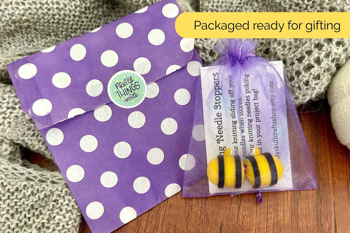 A gift set featuring purple and white polka dot paper envelopes and a sheer purple gift bag containing two bumblebee-shaped needle stoppers, styled on a wooden surface with a knitted blanket background.