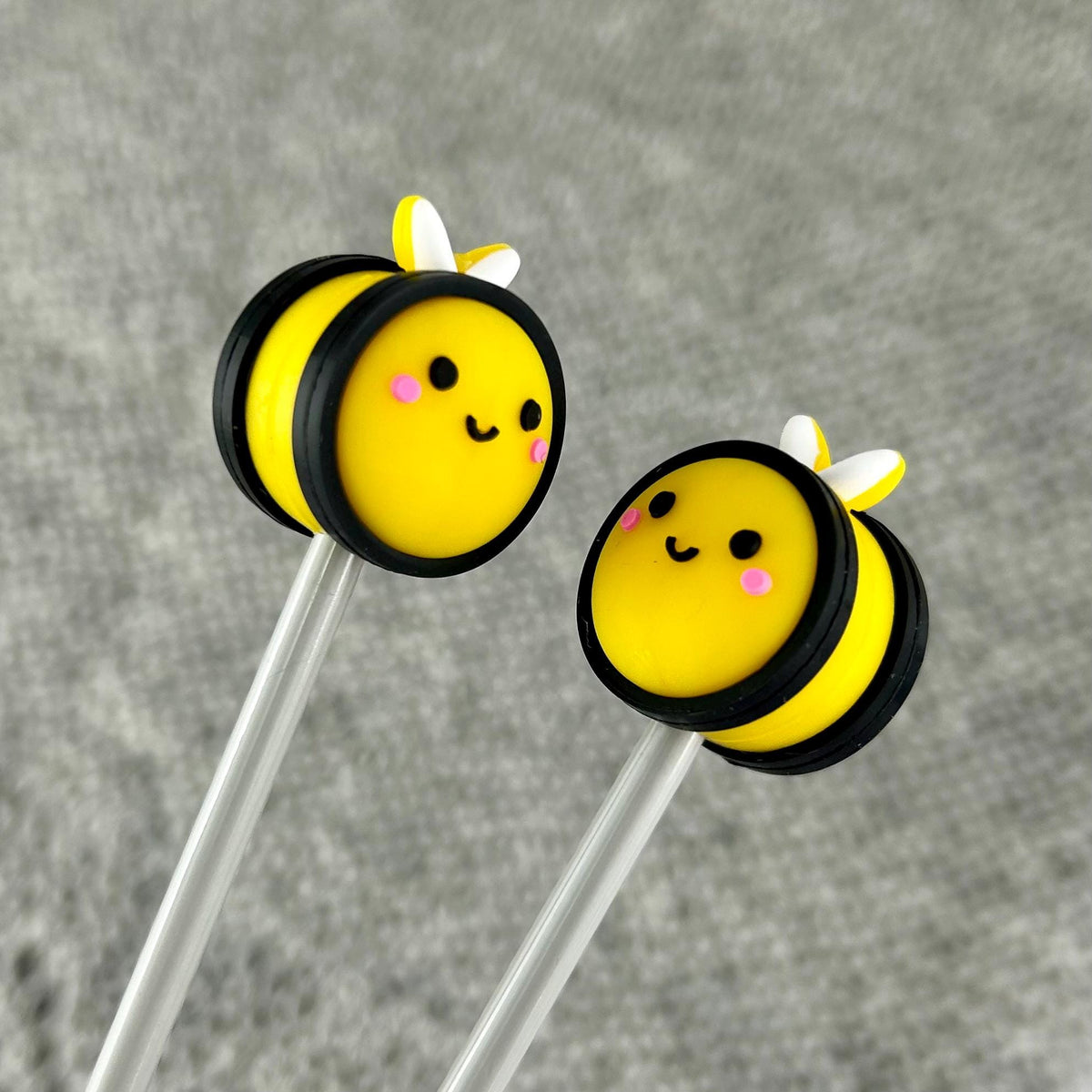 Two yellow and black bee-shaped knitting needle stoppers with white wings and pink cheeks on metal knitting needles photographed against a textured gray background.