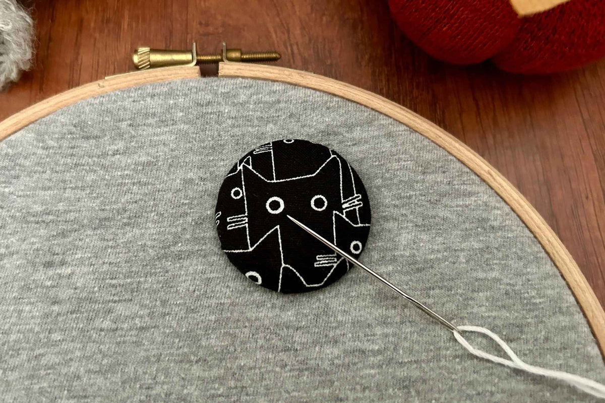 A black cat magnetic needle minder or needle nanny rests on a grey fabric in a wooden embroidery hoop. A needle with White thread is attached. 