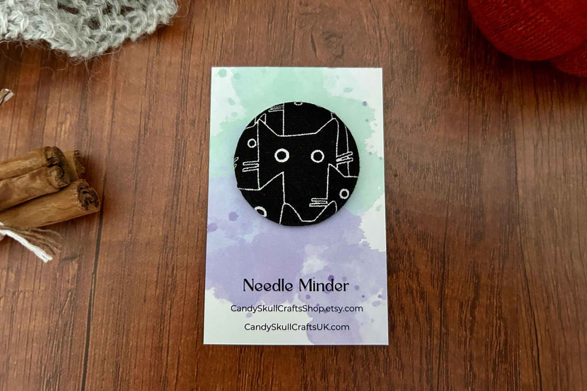 A round black cat with a white outline magnetic needle minder on a printed backing card labeled 'Needle Minder' and 'Candy Skull Crafts', placed on a wooden surface with knitting and yarn props around it.
