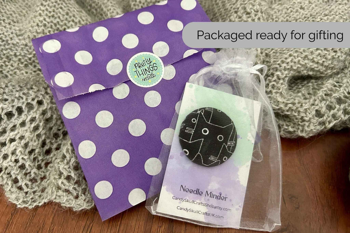A packaged black cat magnetic needle minder packaged in an organza bag next to the purple and white polka dot bag it comes in. 