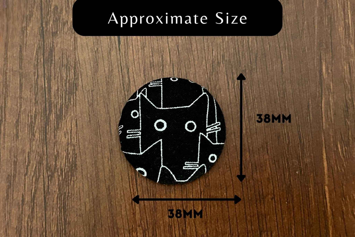 A circular black cat magnetic needle minder with a white line-art cat face sits on a brown wood background. A black label at the top reads 'Approximate Size' with arrows indicating the needle nanny is about 38mm by 38mm.