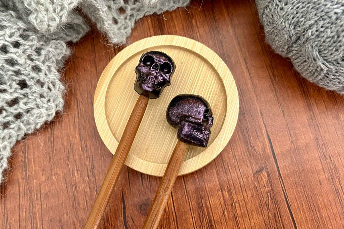 Two with dark purple skull-shaped needle stoppers on the tips resting on a round bamboo coaster, set on a wooden table with gray knitted fabric in the background.