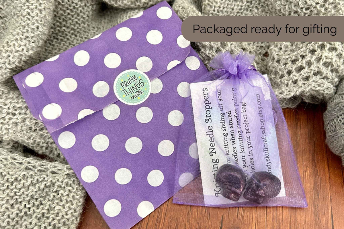 Purple polka dot gift packaging with a matching envelope and a small sheer purple bag containing black skull shaped needle stoppers, styled on a wooden surface with a knitted blanket. A label reads 'Packaged ready for giving' 