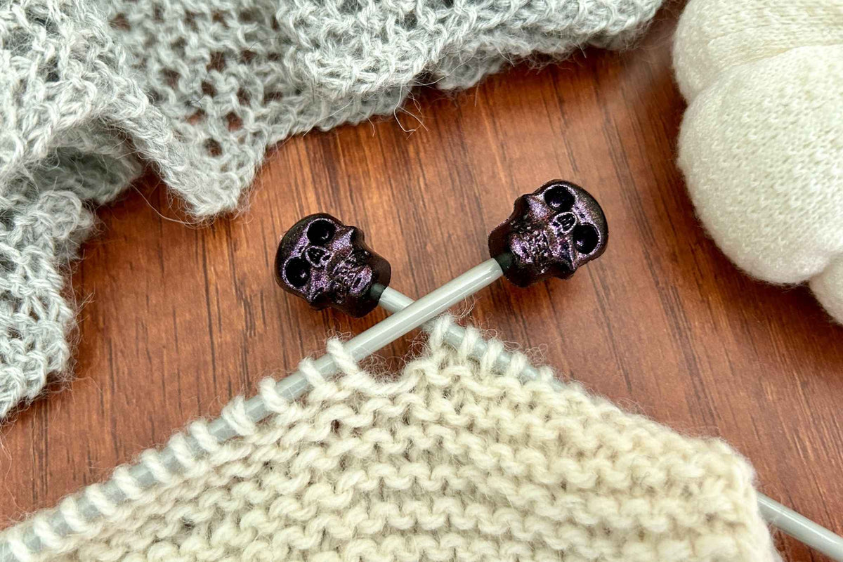 Close-up of knitting needles with skull-shaped black tips, shown stopping cream knitting falling off the ends 