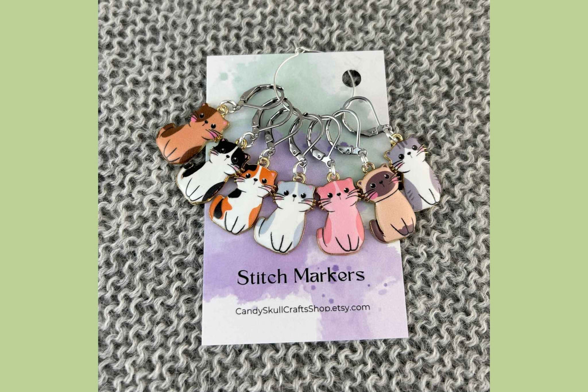 A set of seven knitting or crochet stitch markers featuring cute cartoon cat charms in various colours, attached to a white card labelled 'Stitch Markers' with the brand 'Candy Skull Crafts' visible, set against a gray knitted background.