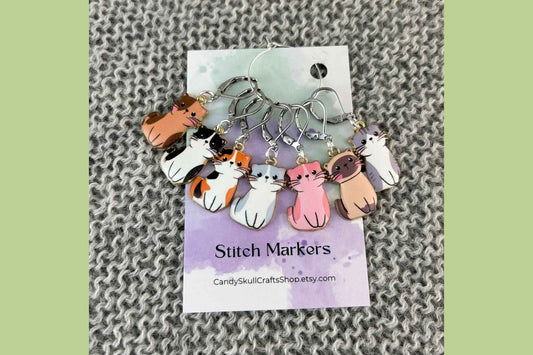 A set of seven knitting or crochet stitch markers featuring cute cartoon cat charms in various colours, attached to a white card labelled 'Stitch Markers' with the brand 'Candy Skull Crafts' visible, set against a gray knitted background.