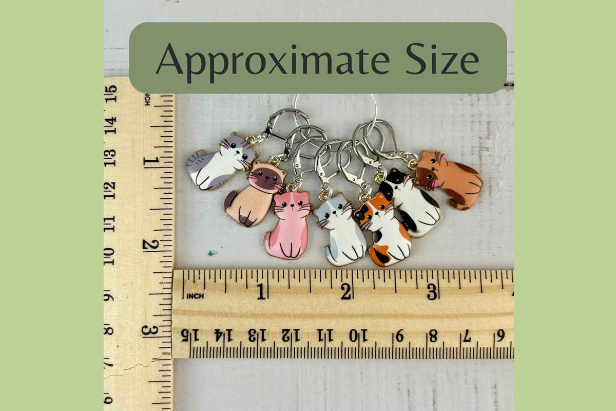 Six small cat-shaped knitting stitch markers arranged above two wooden rulers with an 'Approximate Size' label, showing the scale of the items. Each charm is approx 1inch 