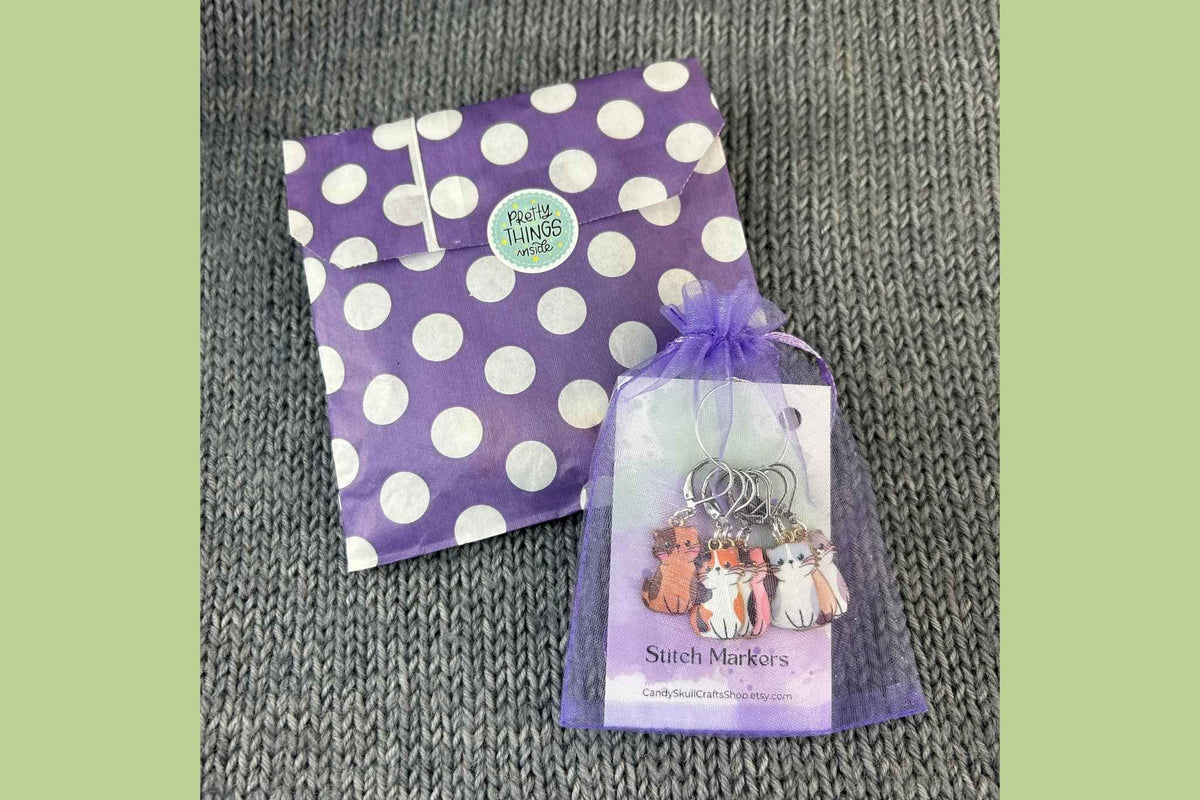 A small gift set featuring a purple and white polka dot envelope and a sheer purple bag holding seven cat knitting stitch markers, displayed on a gray textured fabric background.