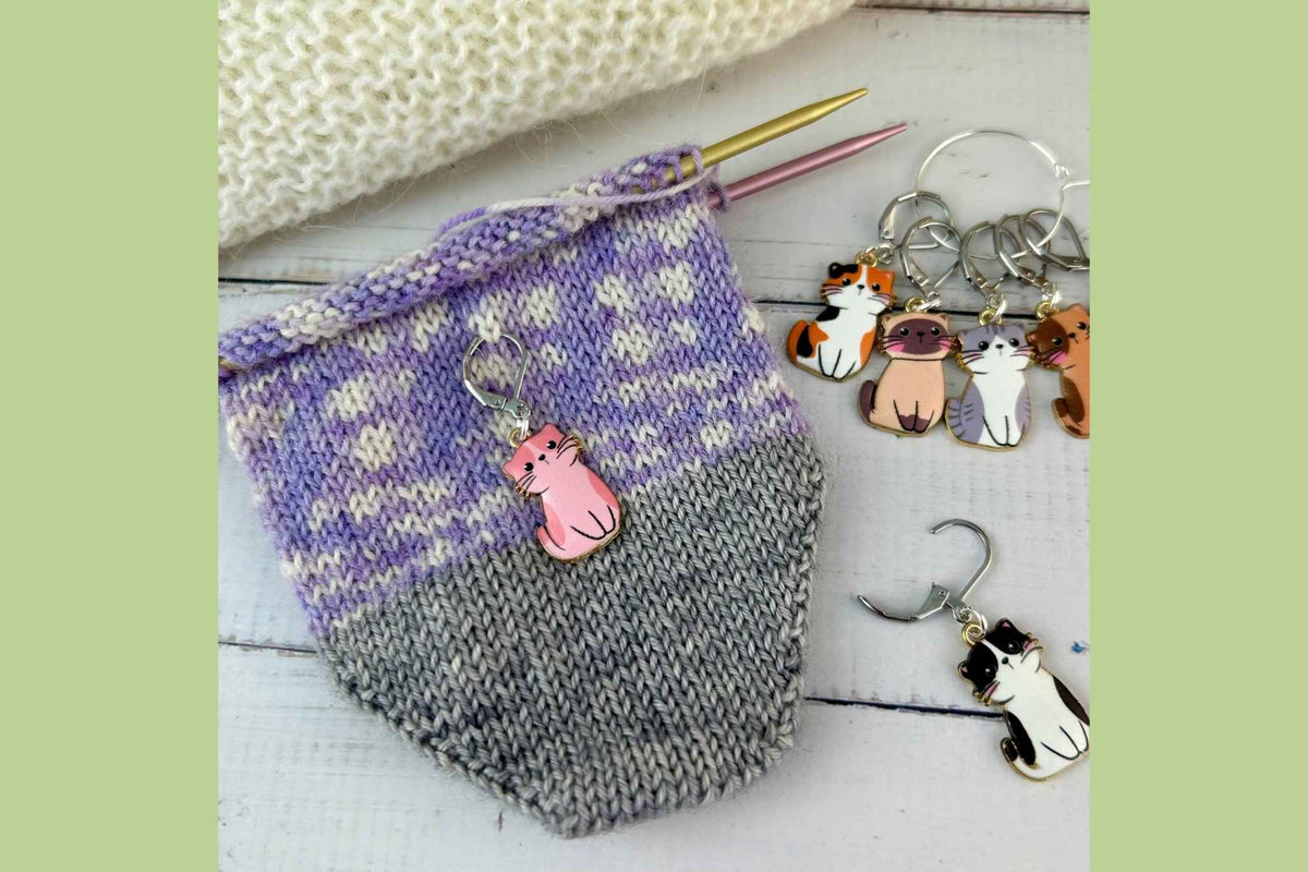 A knitted knitting sock with a purple and gray pattern and a pink cat knitting stitch marker and progress keeper set, shown alongside a white knitted blanket on a light wood surface.