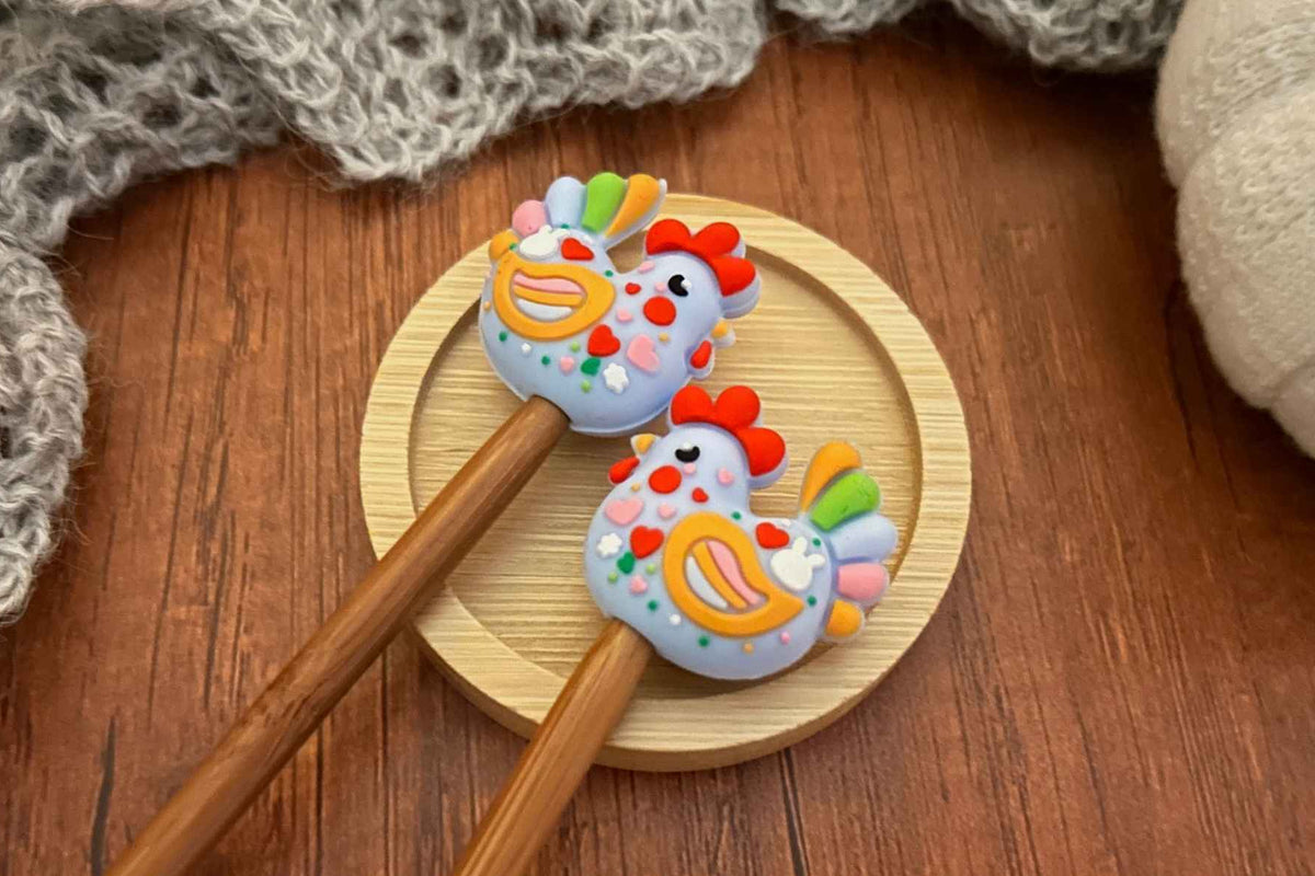 two chicken shaped needle stitch savers on wooden needles displayed on a small round wooden tray over a brown wood surface with a gray knitted textile in the background.