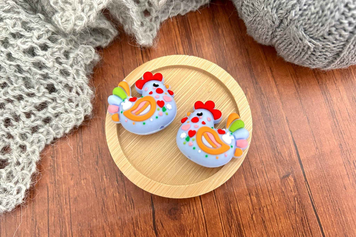 Two small decorative chicken-shaped knitting accessories displayed on a round wooden tray, with a gray knitted blanket on a brown wood surface.