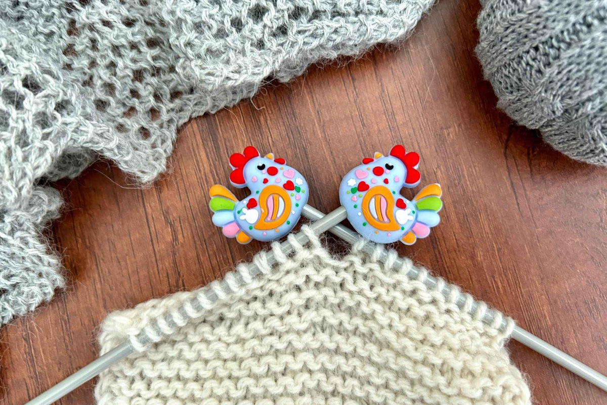 Two colourful chicken knitting needle stoppers resting on cream-colored knitted yarn, with gray yarn and a gray knitted fabric in the background on a wooden surface.