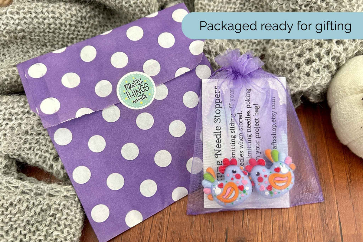 A purple polka dot envelope and a small clear mesh gift bag containing colourful chicken shaped needle stoppers, styled on a wooden surface with a gray knit blanket. 