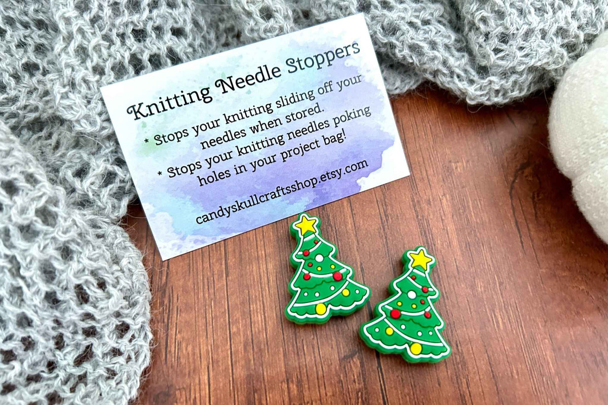 Two small Christmas tree-shaped knitting needle stoppers on a wooden surface next to a product instructions card, styled with a gray knitted scarf.