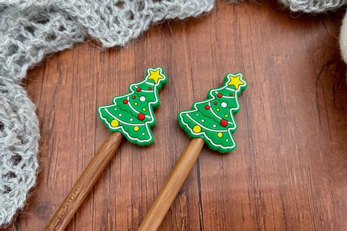 Two wooden knitting needles with green Christmas tree-shaped needle stoppers decorated with yellow, red, and white accents, resting on a wooden surface with a gray knitted scarf in the background.