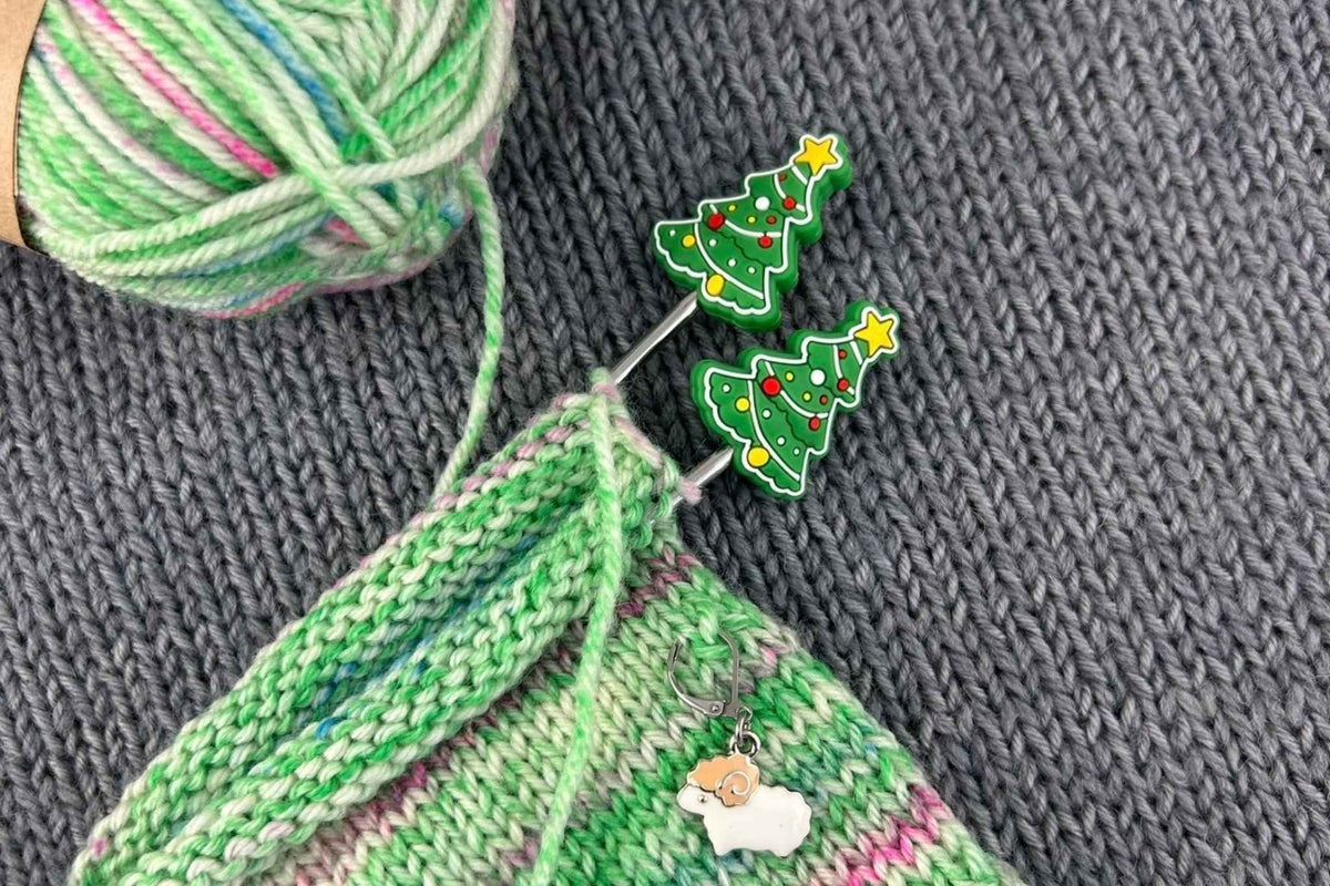 Two Christmas tree-shaped metal knitting pins with colorful lights and a small sheep charm, shown inserted into a green and pink knitted piece next to a ball of variegated yarn on a gray knitted surface.