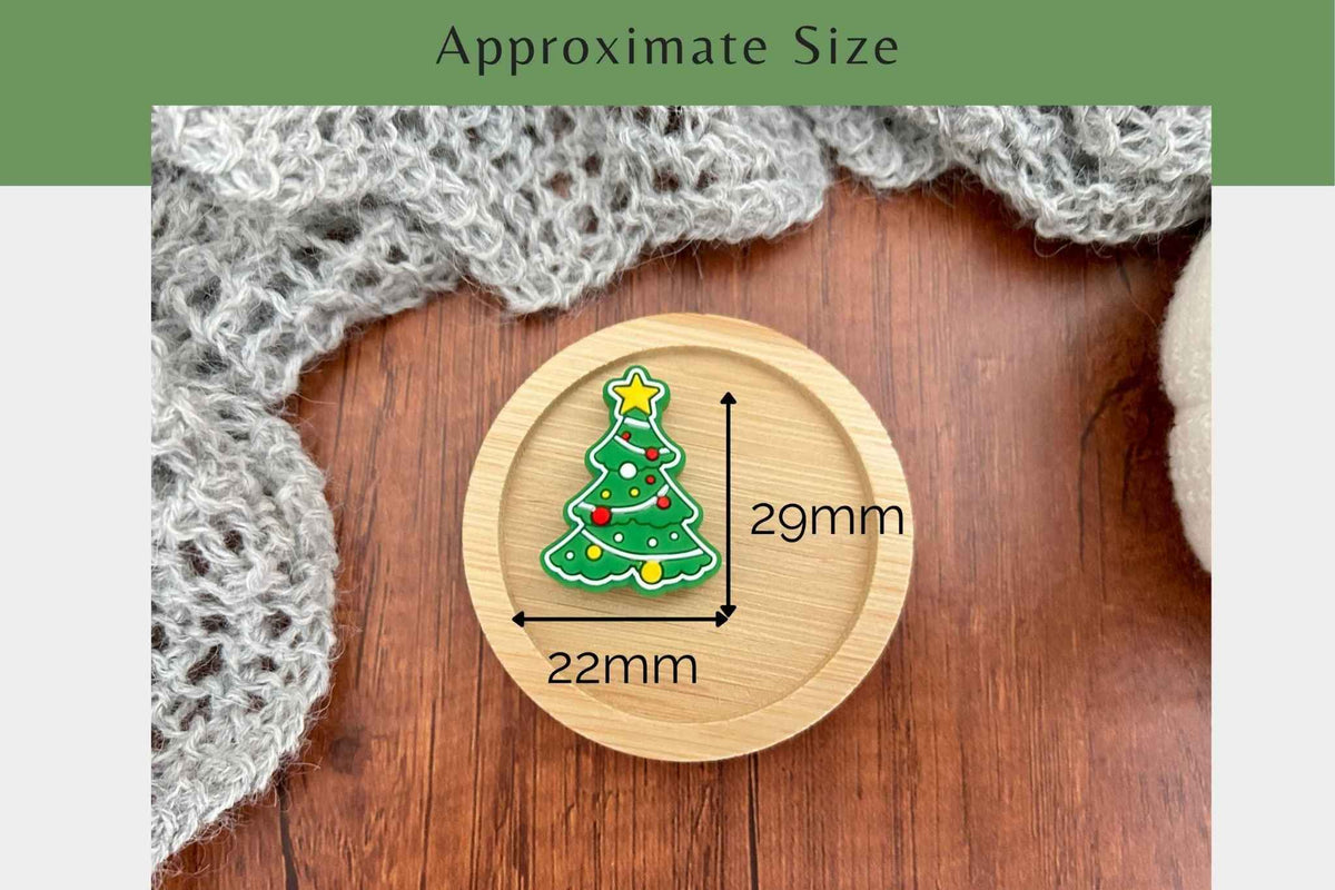 A Christmas tree-shaped needle stopper displayed on a round wooden coaster, with dimension arrows and labels indicating approximate size as 22mm by 29mm. The scene is styled on a wooden surface with a gray knitted scarf, and a green banner at the top reads 'Approximate Size'.