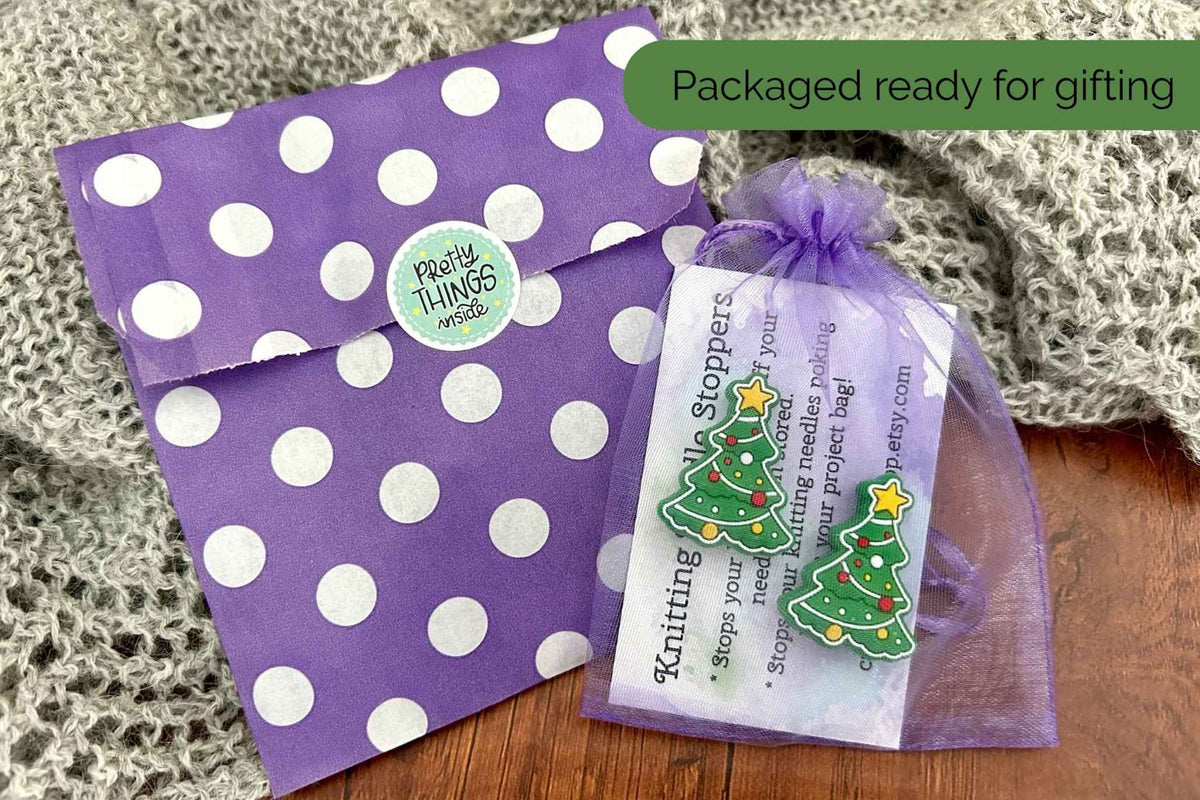 A purple and white polka dot gift pouch with a 'Pretty Things inside' sticker, shown next to a matching purple organza tag bag containing a set of 2 Christmas tree stoppers, on a wooden surface with a gray knit blanket. There is an instruction card in the bag. 