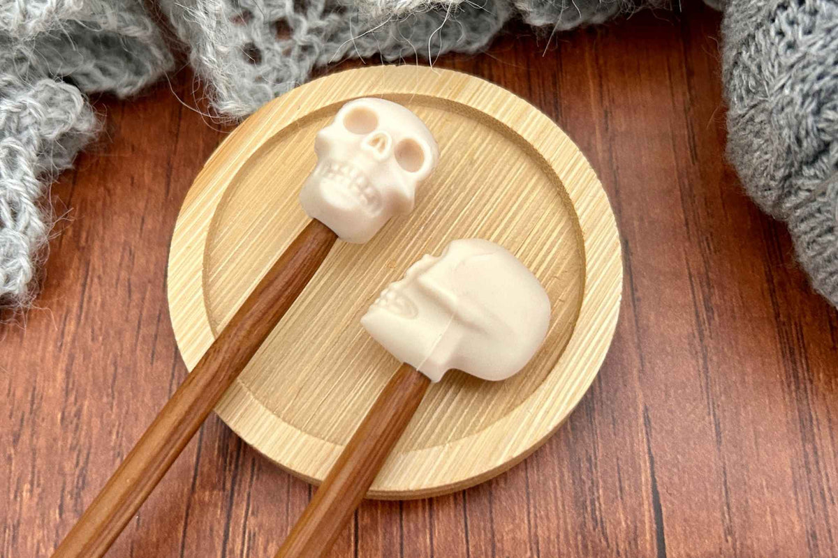 Two cream skull-shaped needle stitch savers on wooden needles displayed on a small round wooden tray over a brown wood surface with a gray knitted textile in the background.
