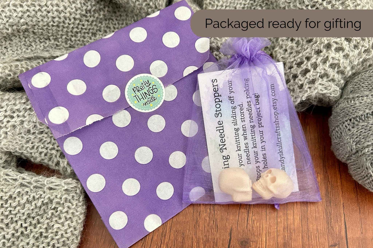 A purple polka dot envelope and a small clear mesh gift bag containing skull shaped shaped stitch savers or stoppers with an instruction card, styled on a wooden surface with a gray knit blanket. 