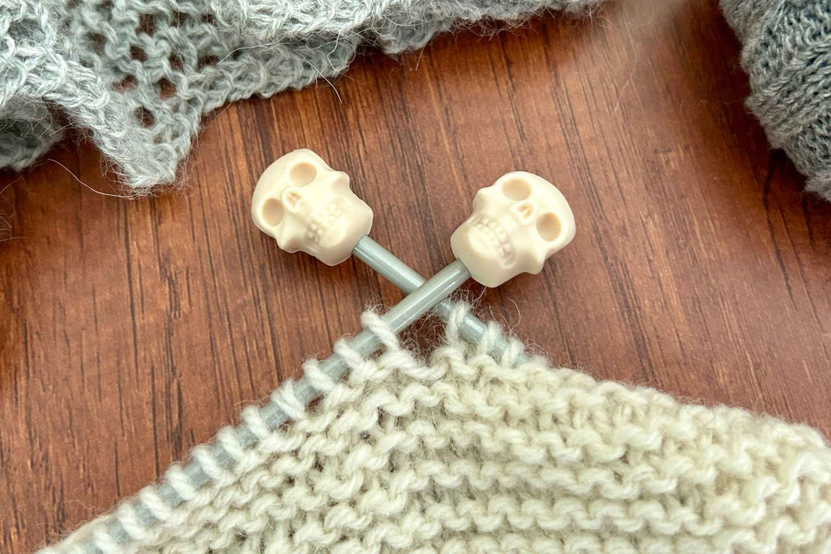 Two cream skull-shaped knitting needle stoppers stopping stitches falling off cream knitting, with a gray knitted fabric in the background on a wooden surface.
nearby.