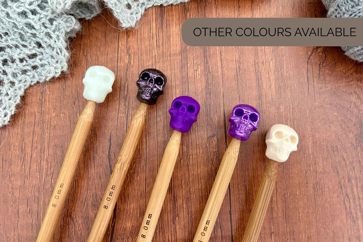 Five wooden knitting needles with skull-shaped handles in white, black, purple, glitter purple and cream, arranged on a wooden surface next to a gray knitted cloth. A text overlay at the top right reads 'OTHER COLOURS AVAILABLE'.