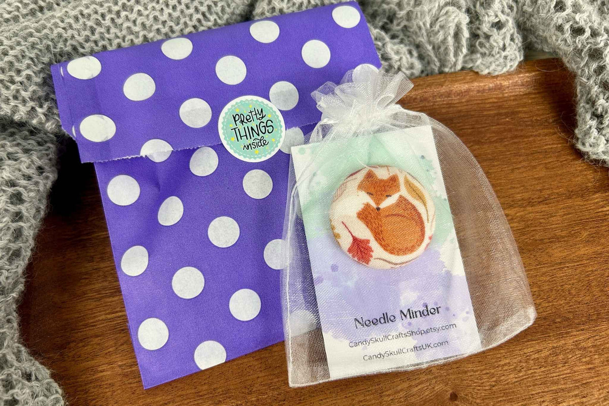 A fox fabric needle minder in a mesh organza bag with a purple and white polka dot envelope next to it. The envelope features a round sticker that reads 'pretty Things inside'.