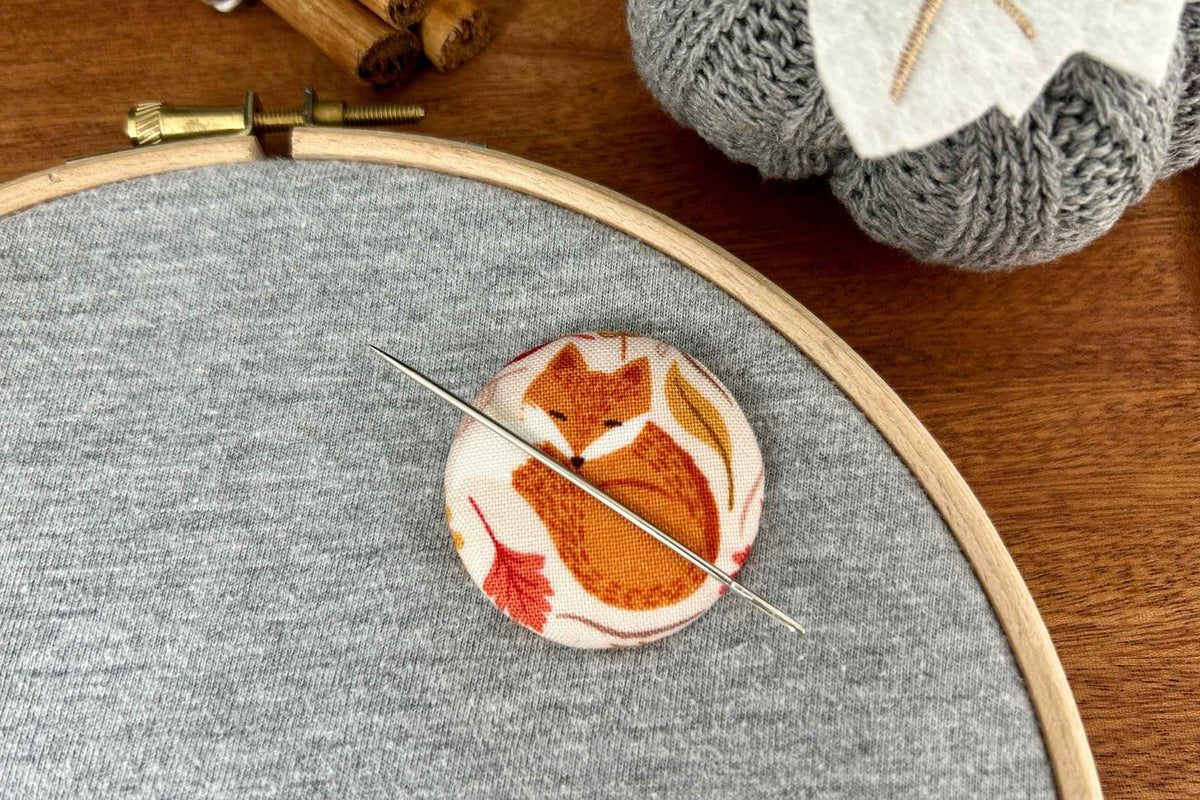 A round magnetic fabric needle minder featuring an orange fox on a cream background, resting on gray fabric inside a wooden embroidery hoop with a sewing needle on top. A gray knitted pumpkin is visible in the background on a wooden table.